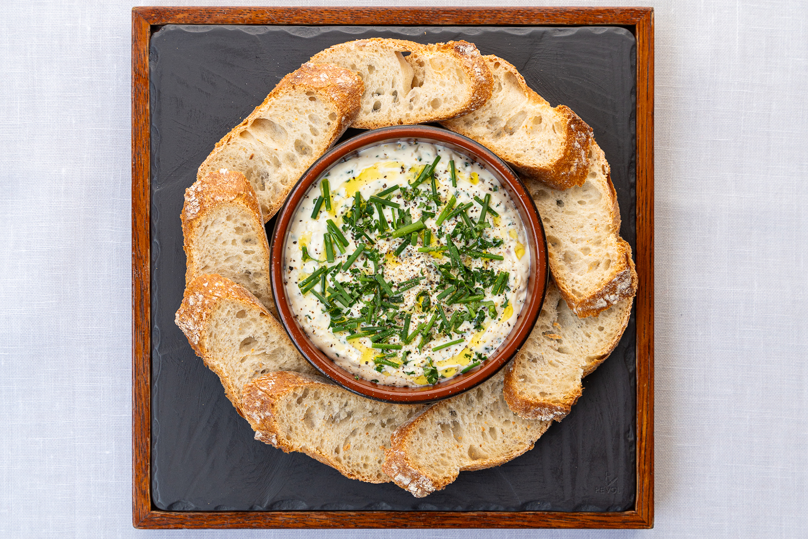 La cervelle des canuts (fromage frais lyonnais servi agrémenté de ciboulette, ail, persil et huile), accompagnée de tranches de baguette de campagne. Ce fromage est servi sur une assiettes en ardoise noire et au cadre en bois.