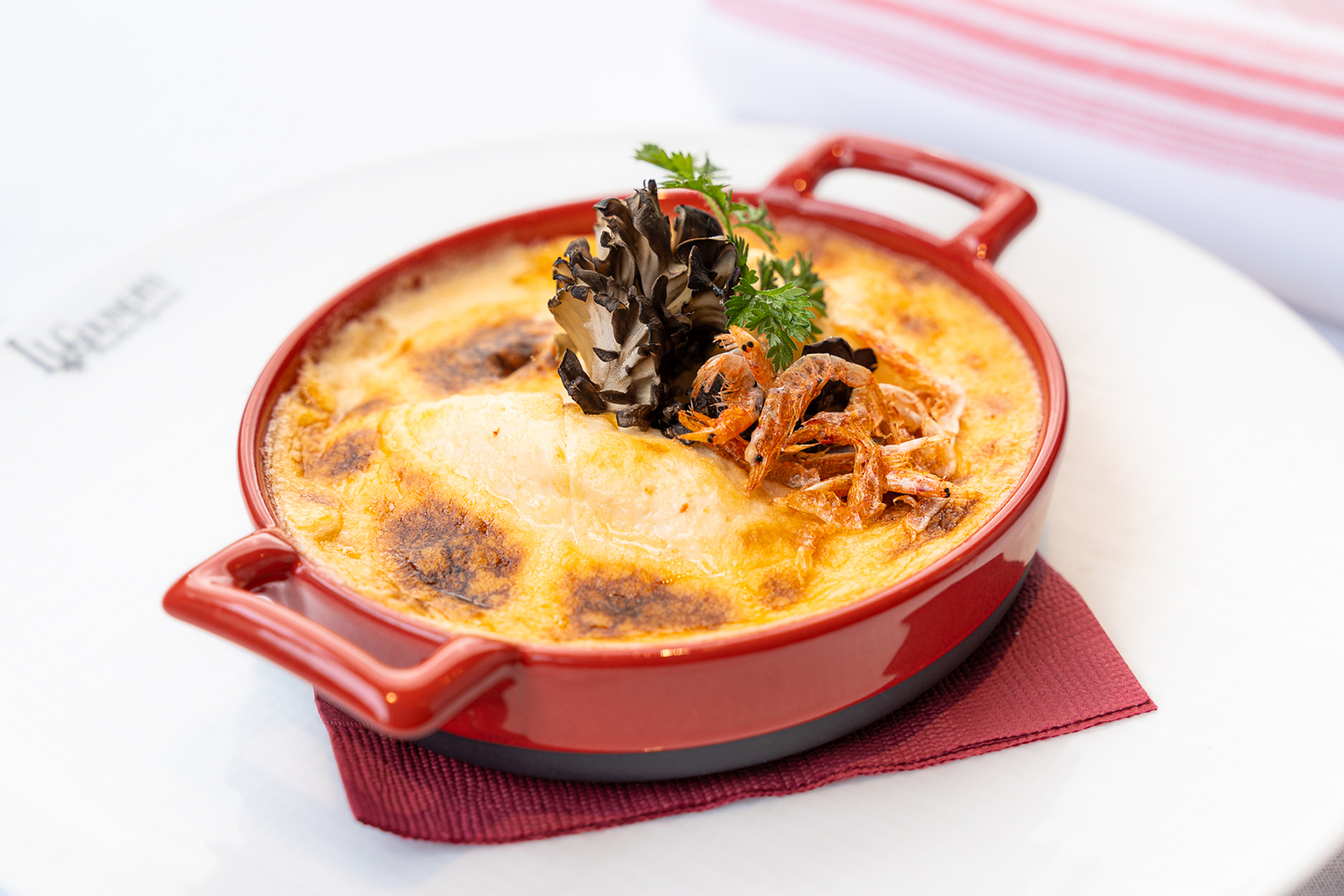 Les fameuses quenelles lyonnaises de la mère Mauricette, servies avec une sauce Nantua. Elles sont présentées dans un plat à four rouge et décorées de quelques petites crevettes grillées, de champignons et d'herbes.