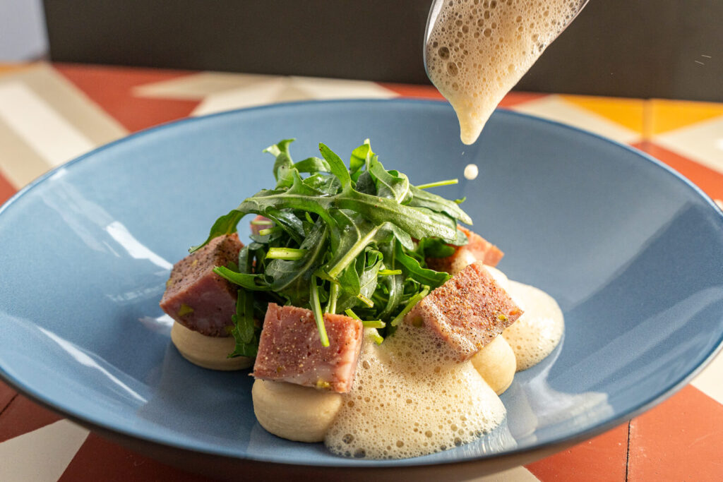 Les morceaux de saucisson lyonnais pistaché sont disposés en rond dans l'assiette et déposés sur la purée de coco pochée. La salade de roquette est placée au centre de l'assiette.