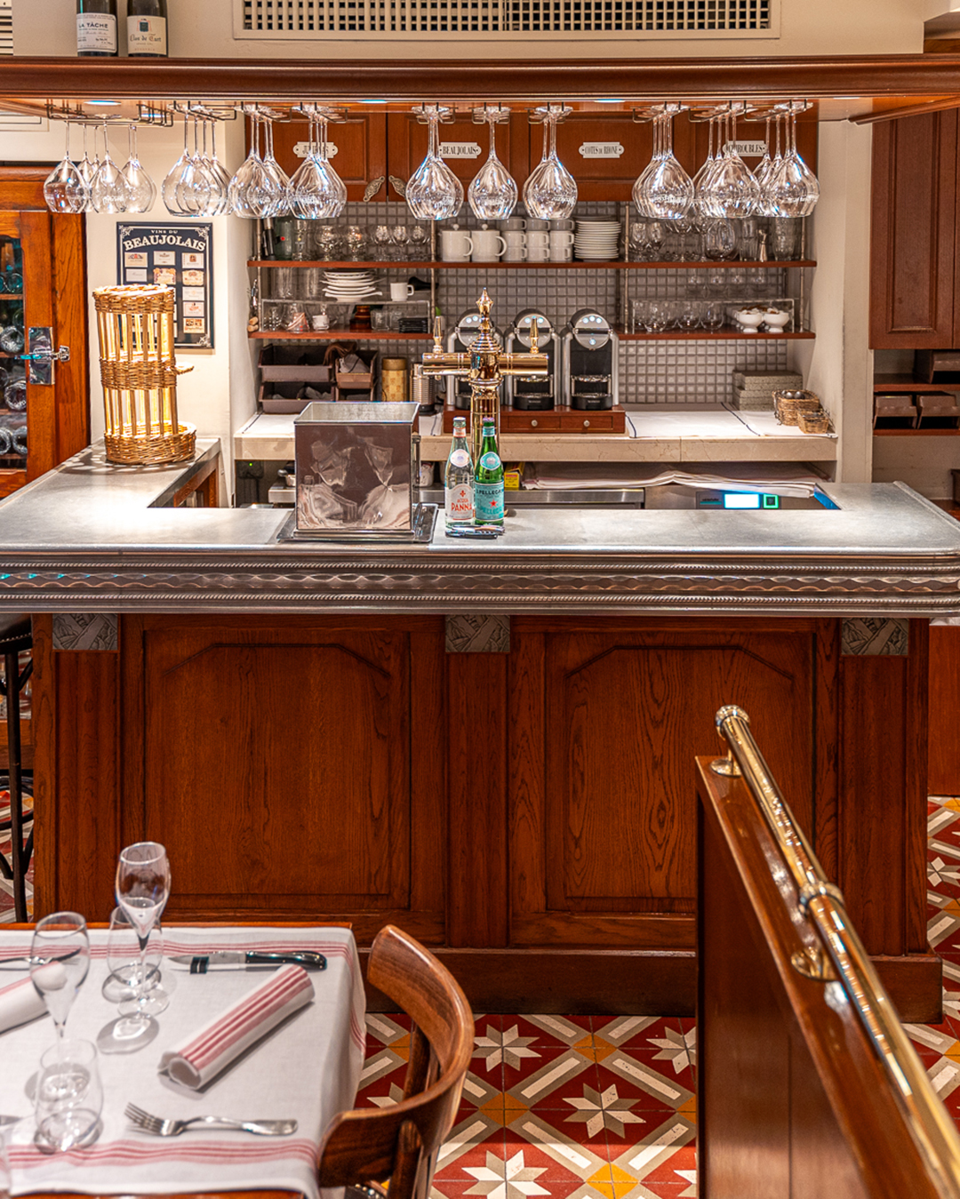 Le bar comptoir en bois et en étain traditionnel des bouchons lyonnais du restaurant Lugdunum Bouchon Lyonnais. On y voit des verres à vin suspendus et une cave à vin sur le côté gauche du bar. Les couleurs brunes et l'atmosphère qui se dégagent de la photo rappellent vraiment celle des bouchons lyonnais en France.
