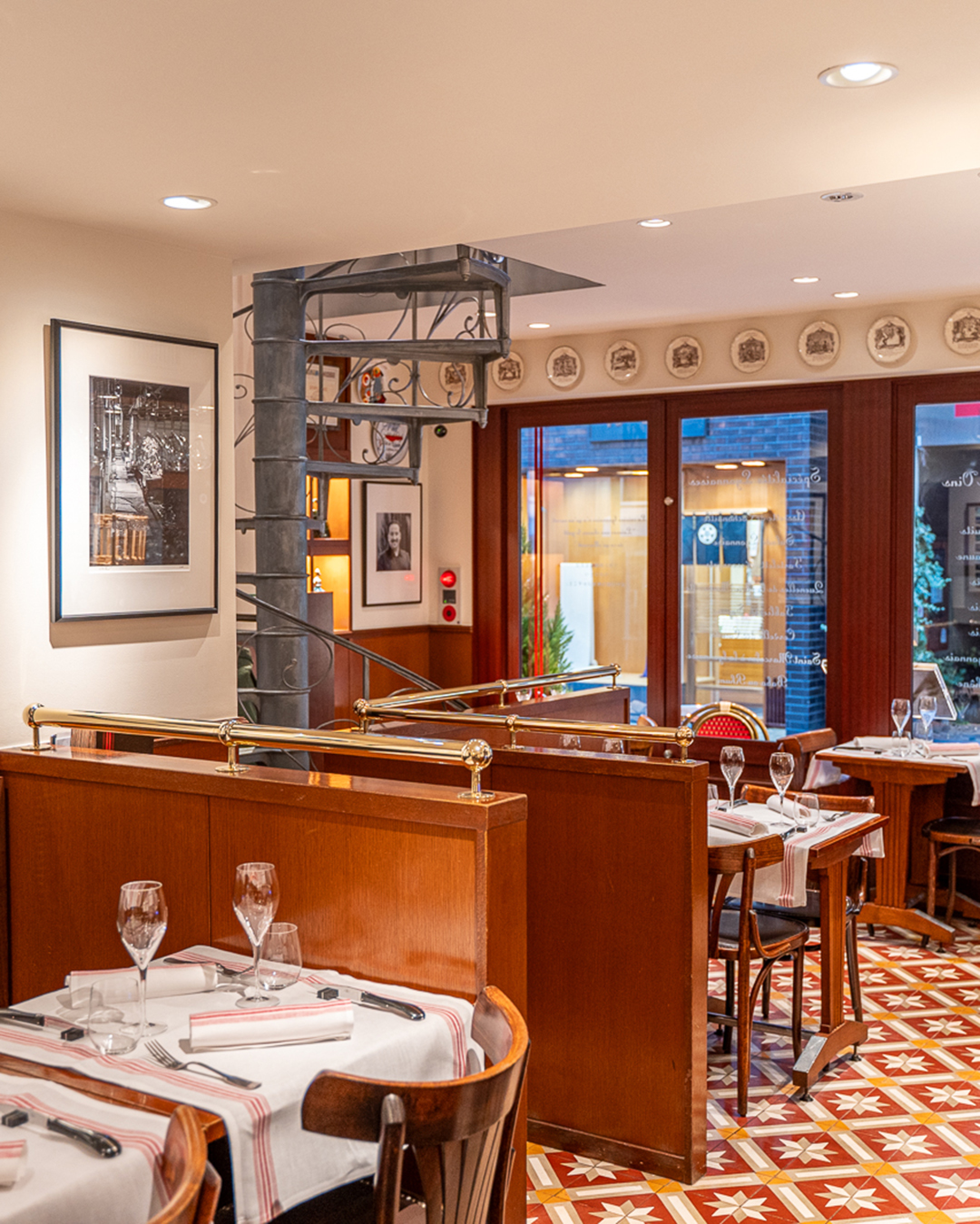 La salle principale du restaurant Lugdunum Bouchon Lyonnais avec les tables dressées, l'escalier en colimaçon de style 1900, et diverses décorations Art Déco qui rappellent la ville de Lyon. À l'arrière-plan, on y voit également les portes vitrées ainsi que la rue.