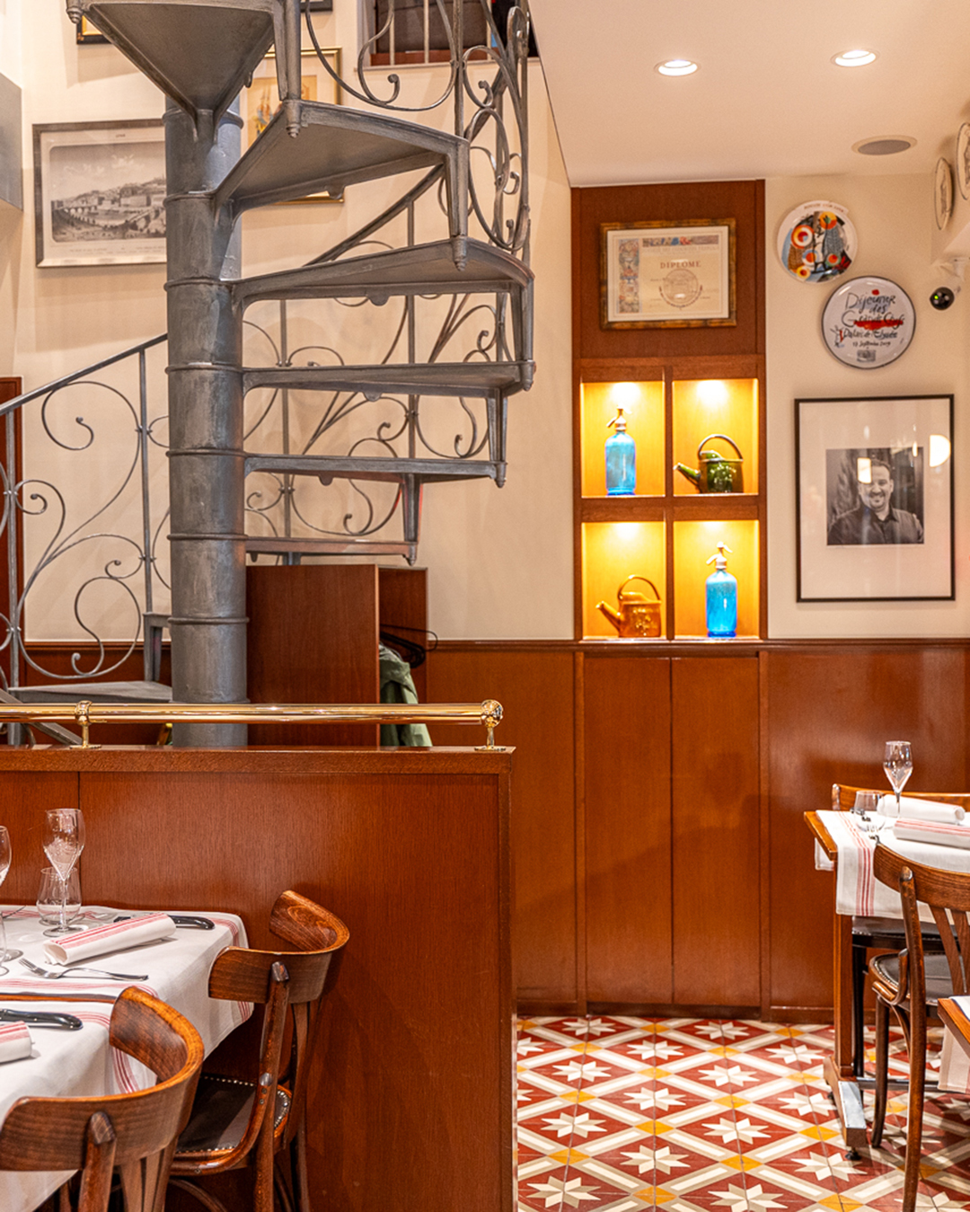 La salle principale du restaurant Lugdunum Bouchon Lyonnais avec les tables dressées, l'escalier colimaçon style 1900 et diverses décorations Art Déco qui rappellent la ville de Lyon.